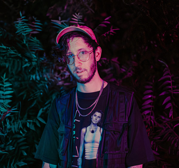 Of The Trees, standing in front of greenery. He is wearing a orange hat and a black shift with necklaces, and there is ambient soft lighting.