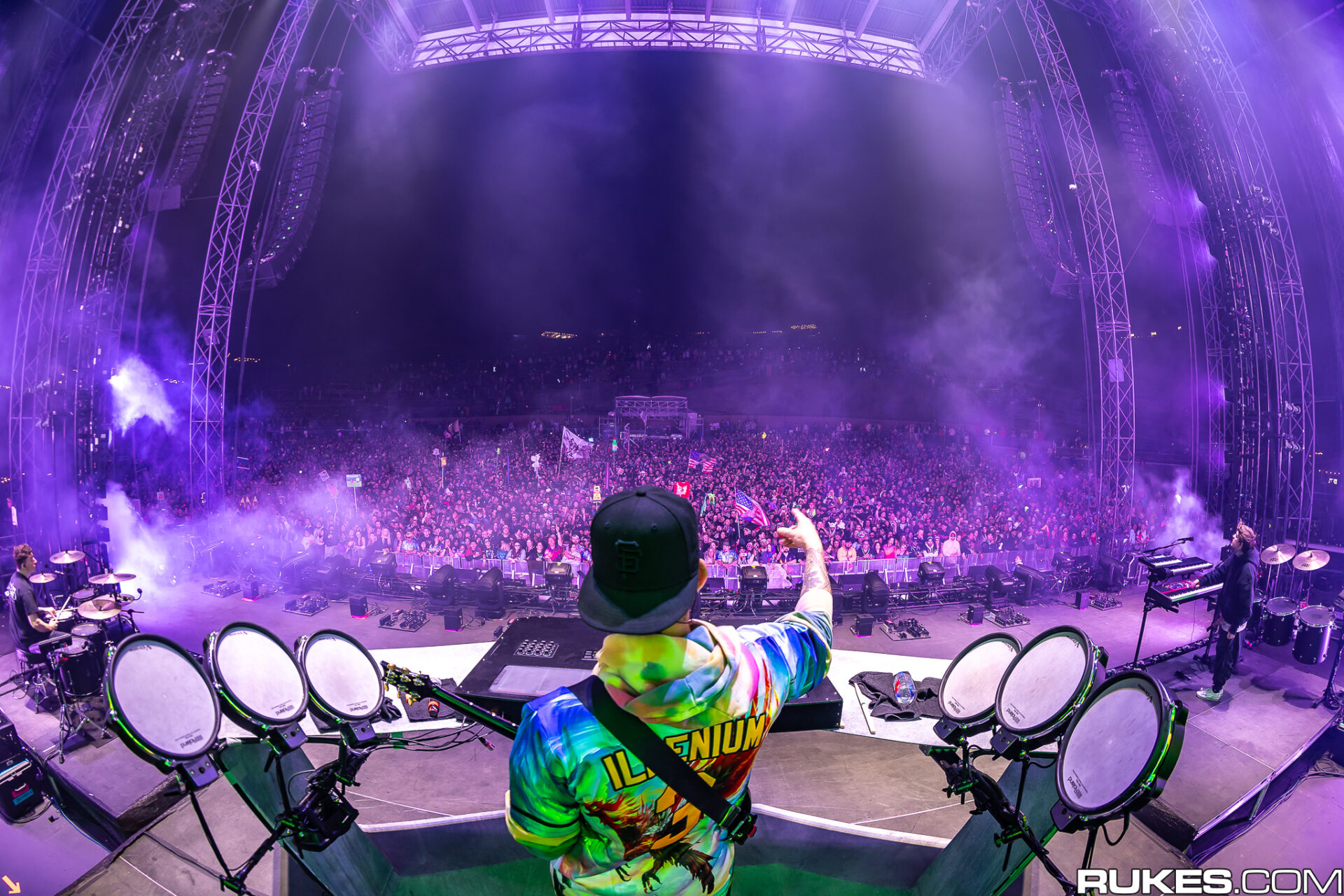 Shot of Illenium performing to pit with DJ set up/drums while holding guitar with purple stage lights. 